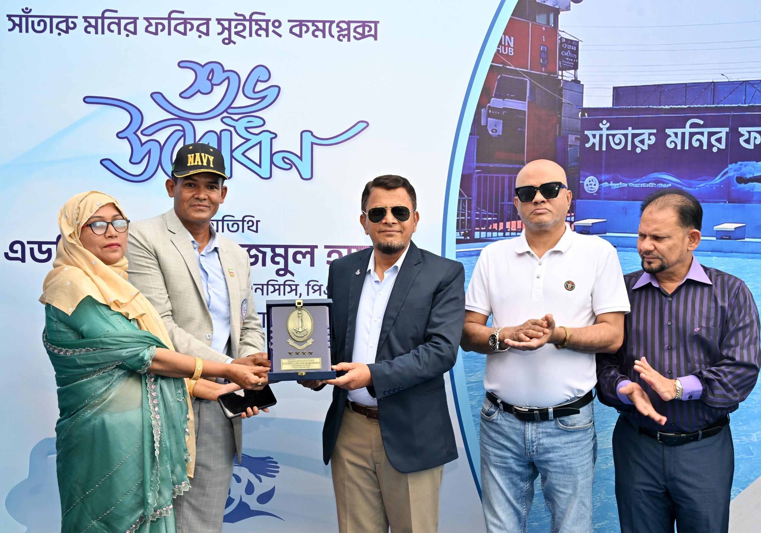 A framed naval crest is presented to a woman in a green saree as officials applaud, the complex's entrance banner behind them.