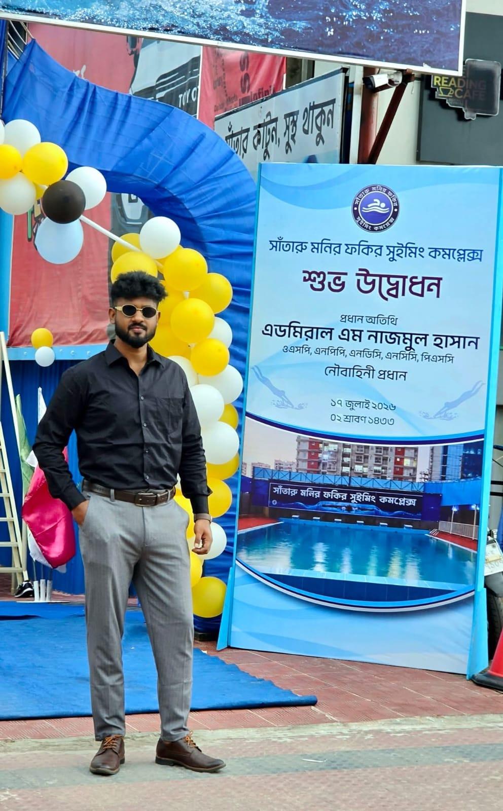 A man in sunglasses poses beside the inauguration signboard and yellow-and-white balloon arch outside Monir Fokir Swimming Complex.