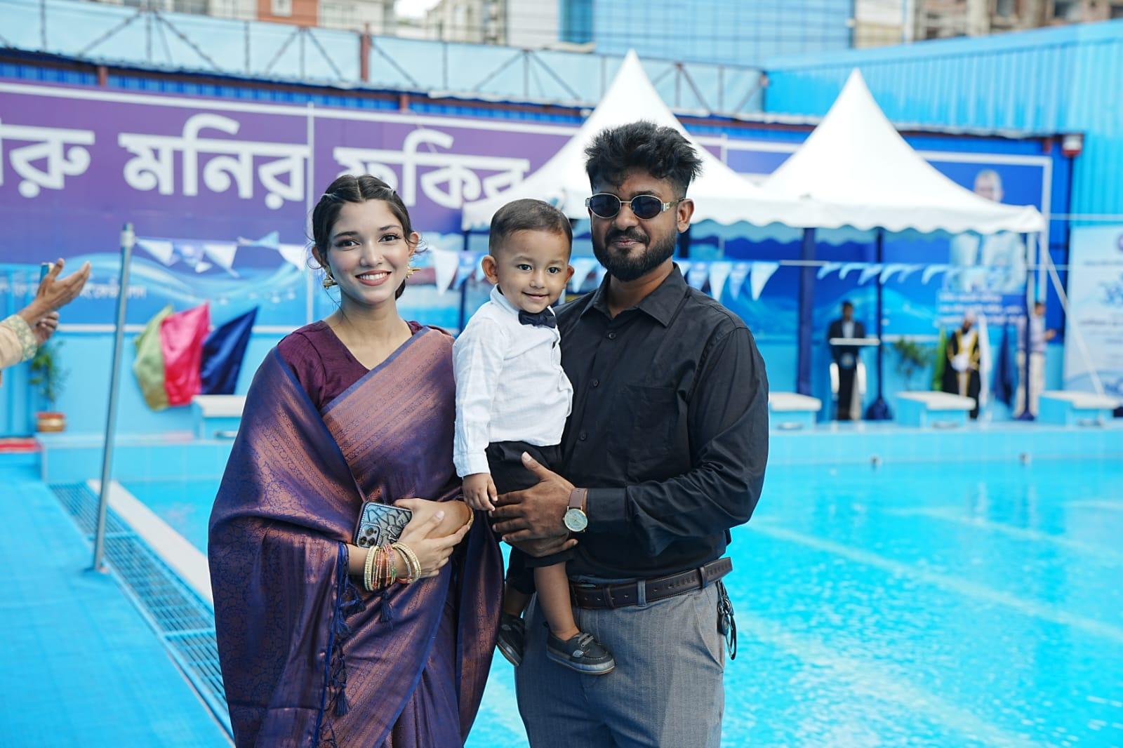 A family poses poolside in formal wear — a woman in a purple saree, a man in sunglasses, and a toddler in a bow tie — with the pool and event tents behind them.