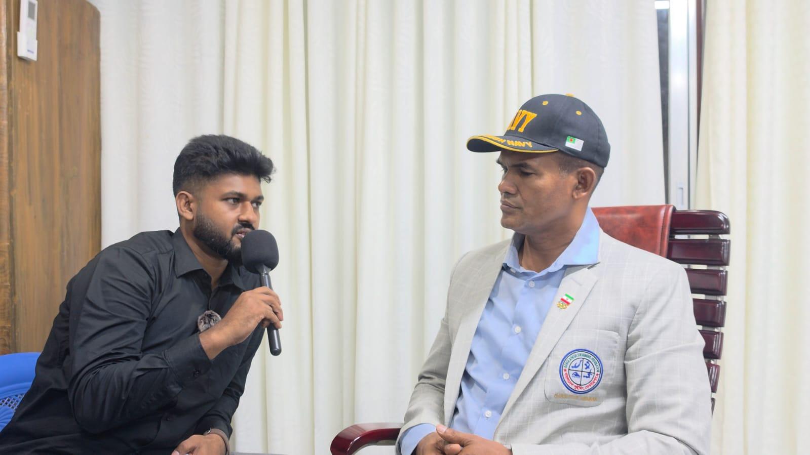 A host holds a microphone toward a Bangladesh Navy officer seated for an interview against a curtained backdrop.