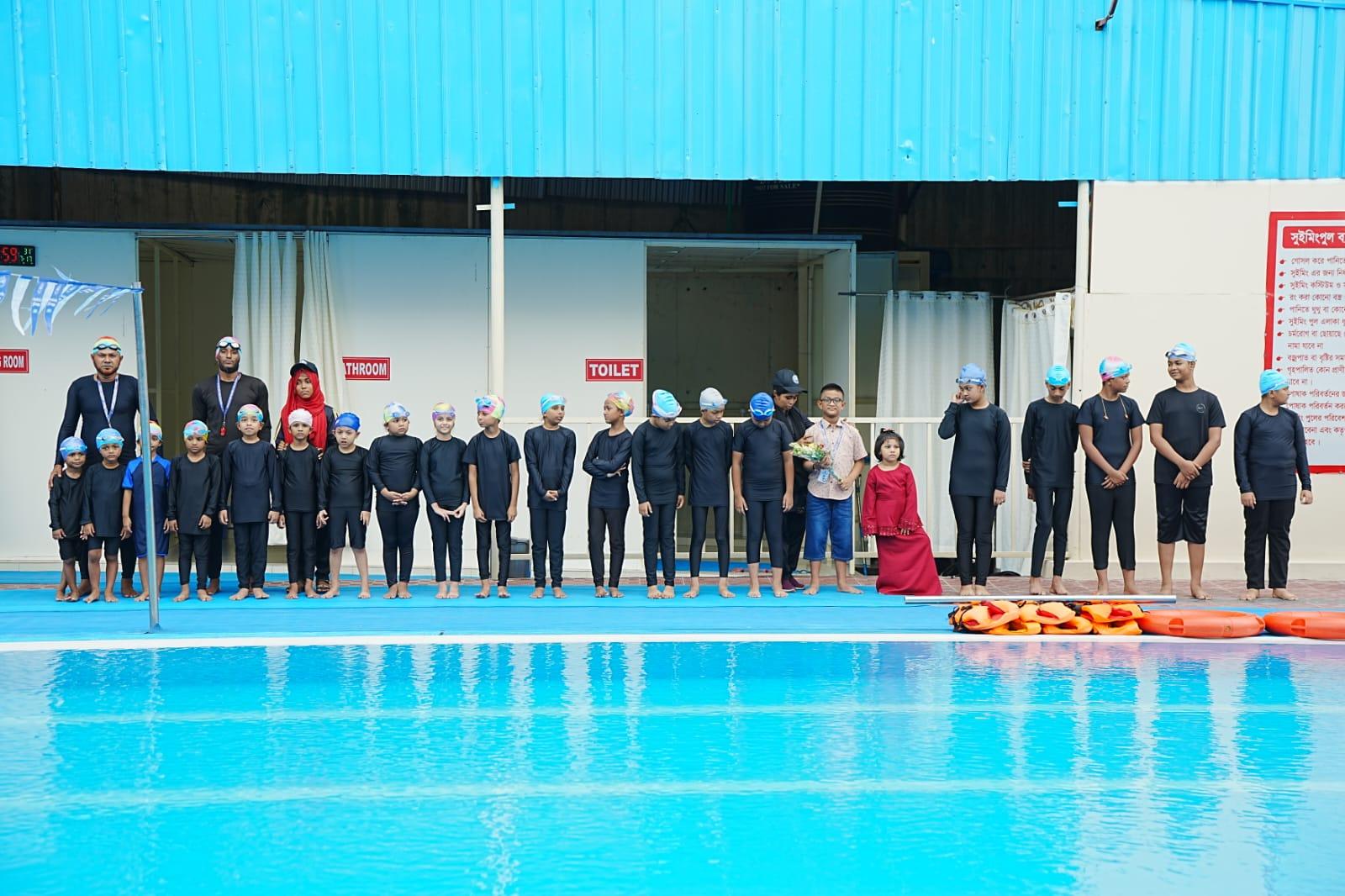 A line of young swimmers in black swimwear and colorful caps stand poolside for a group photo after their demonstration lesson.