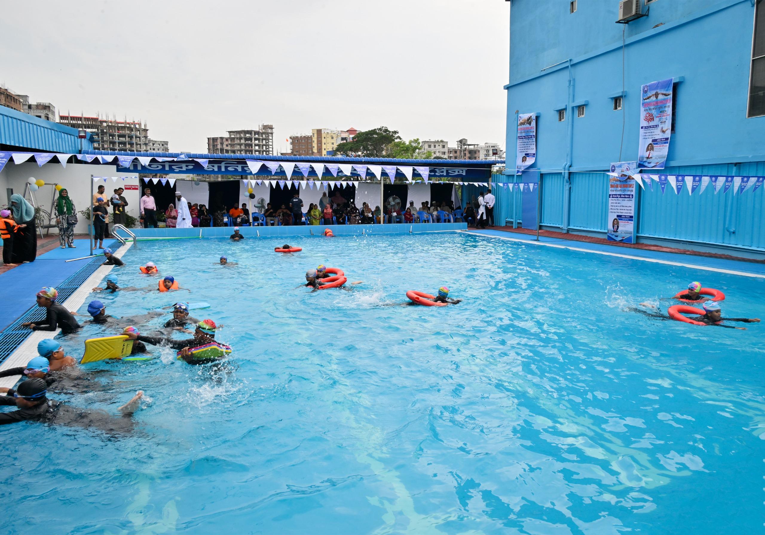 Children in swim caps and life vests practice strokes across the pool as parents and officials look on from poolside chairs.