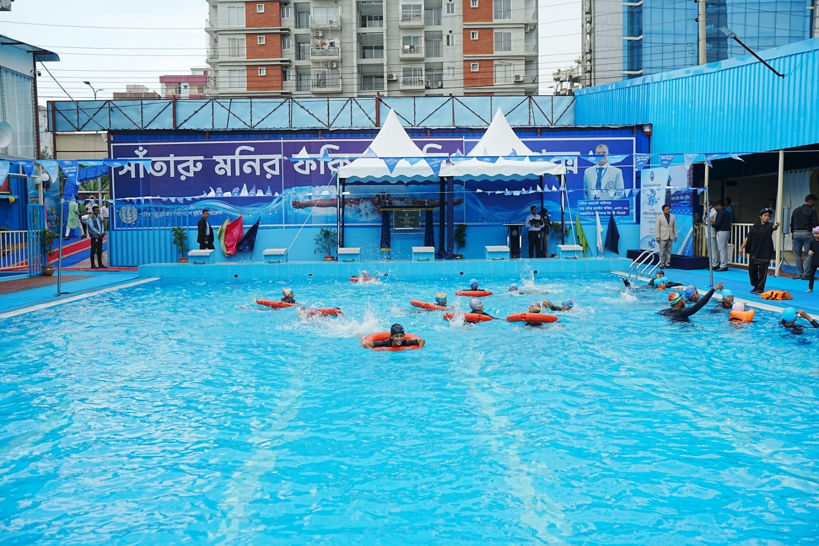 Young swimmers holding orange safety floats swim toward the starting-block platform during the opening-day demonstration.