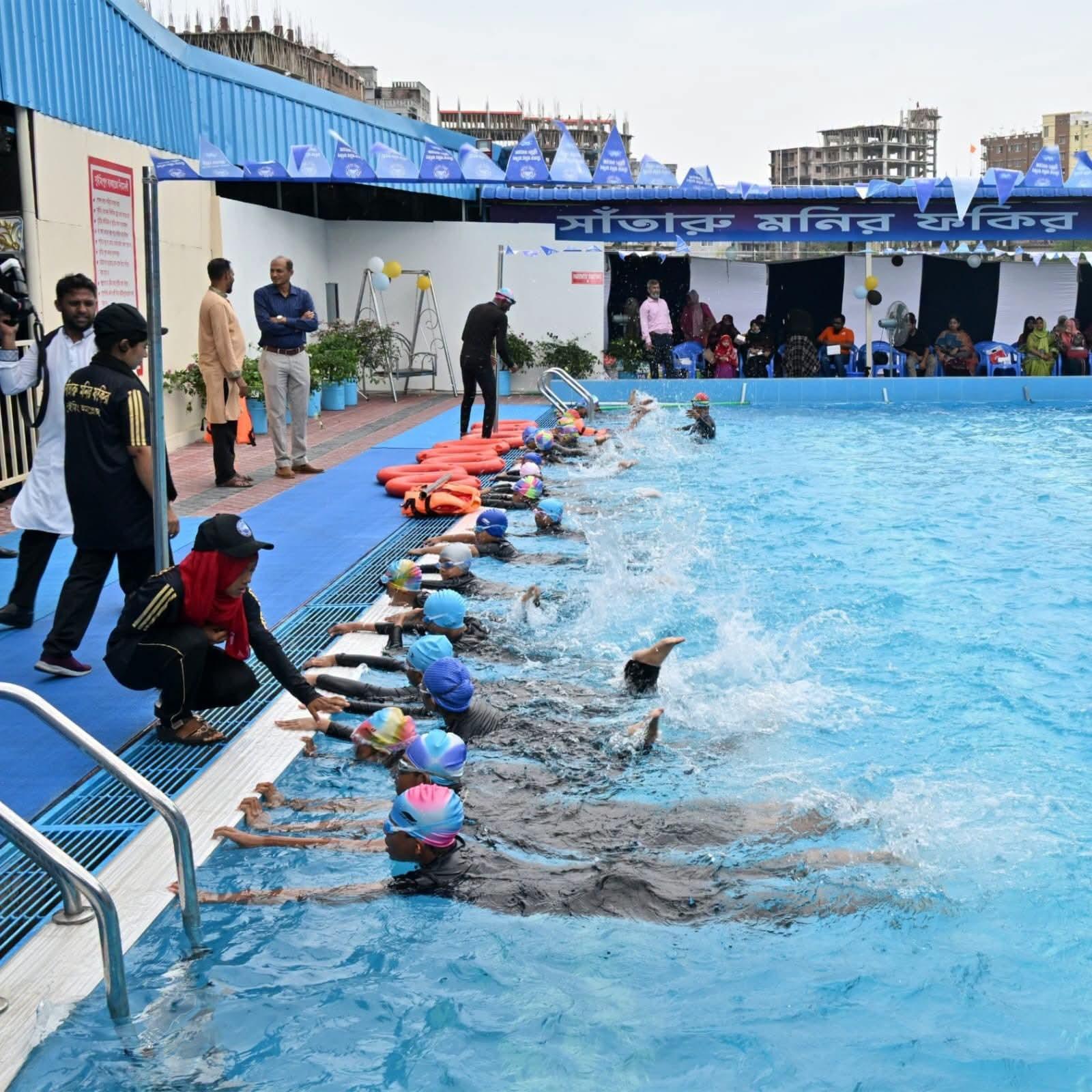 Young swimmers in caps and goggles push off the wall in unison during a demonstration lesson, coaches and seated guests watching from the deck.