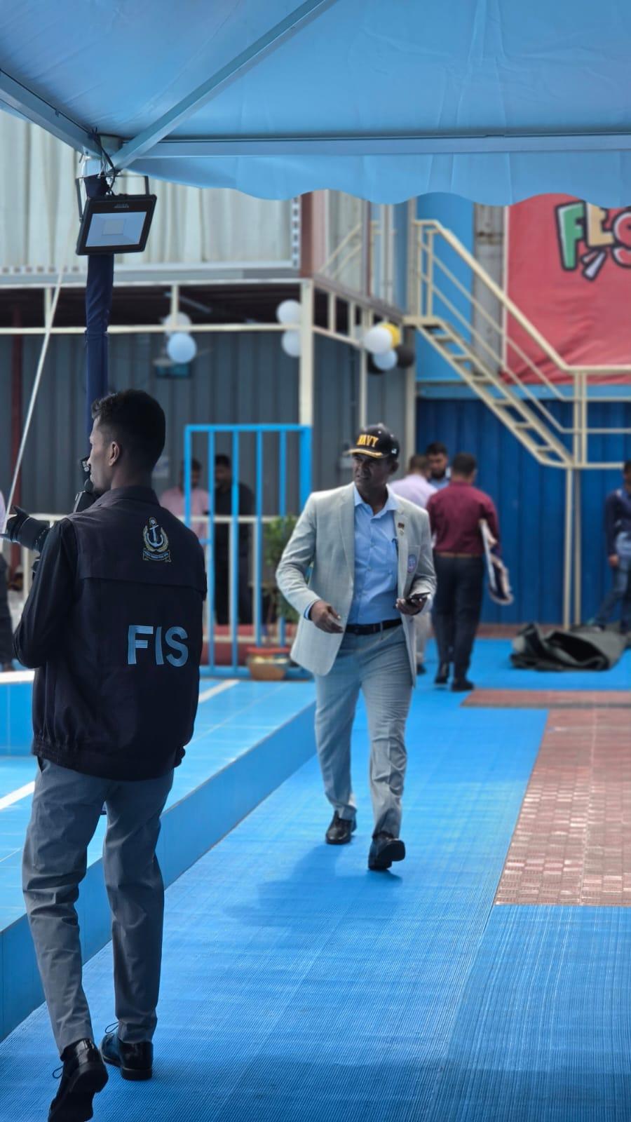 The same Navy officer walks past the starting blocks toward the gathered guests, camera crew following behind.