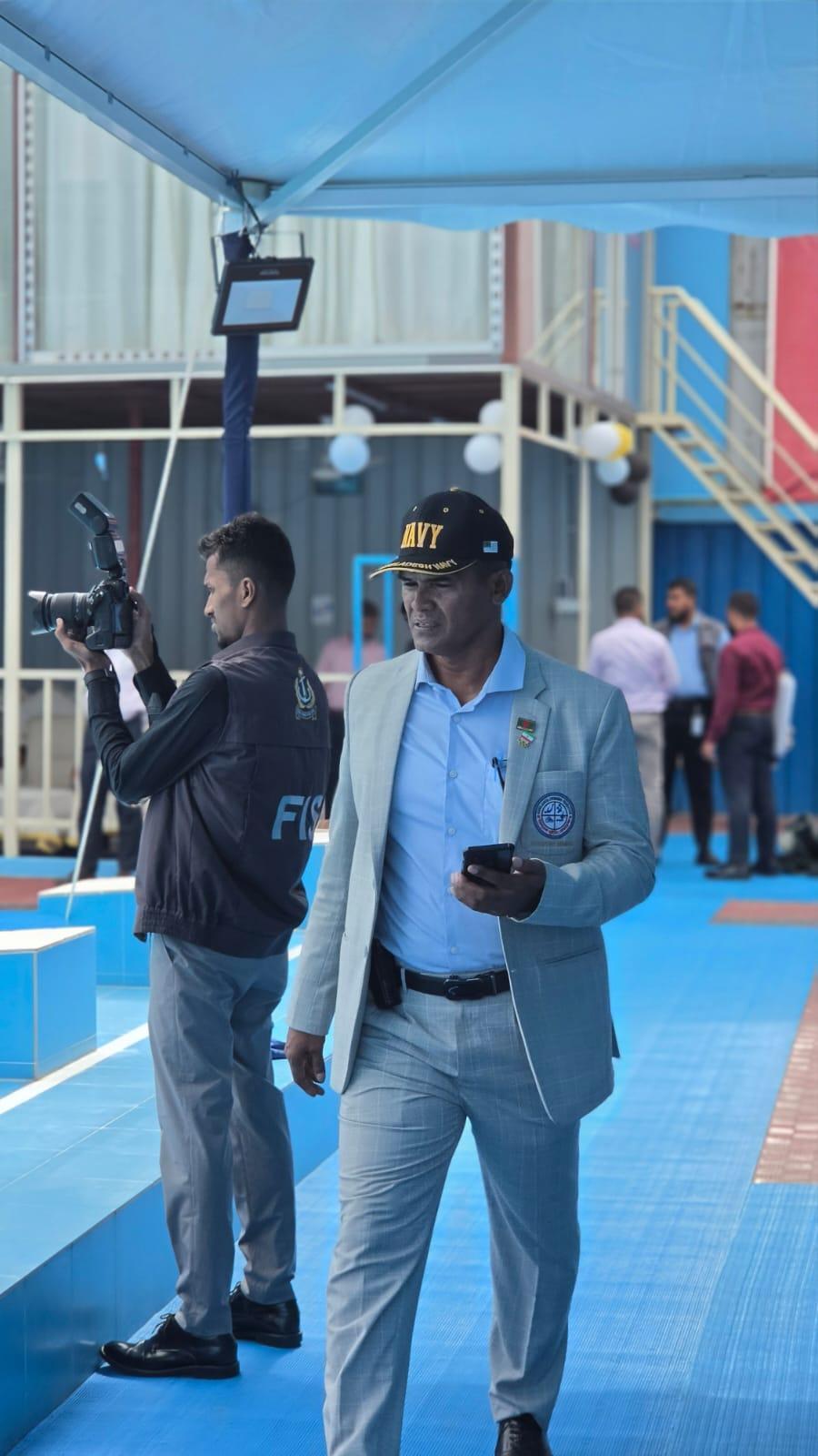 A Bangladesh Navy officer in a cap and light blazer walks the pool deck as a photographer captures the moment.