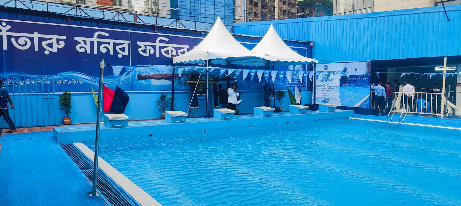 Guests gather near the starting blocks beneath twin canopy tents as the swim complex's banner and mural stretch along the pool wall.