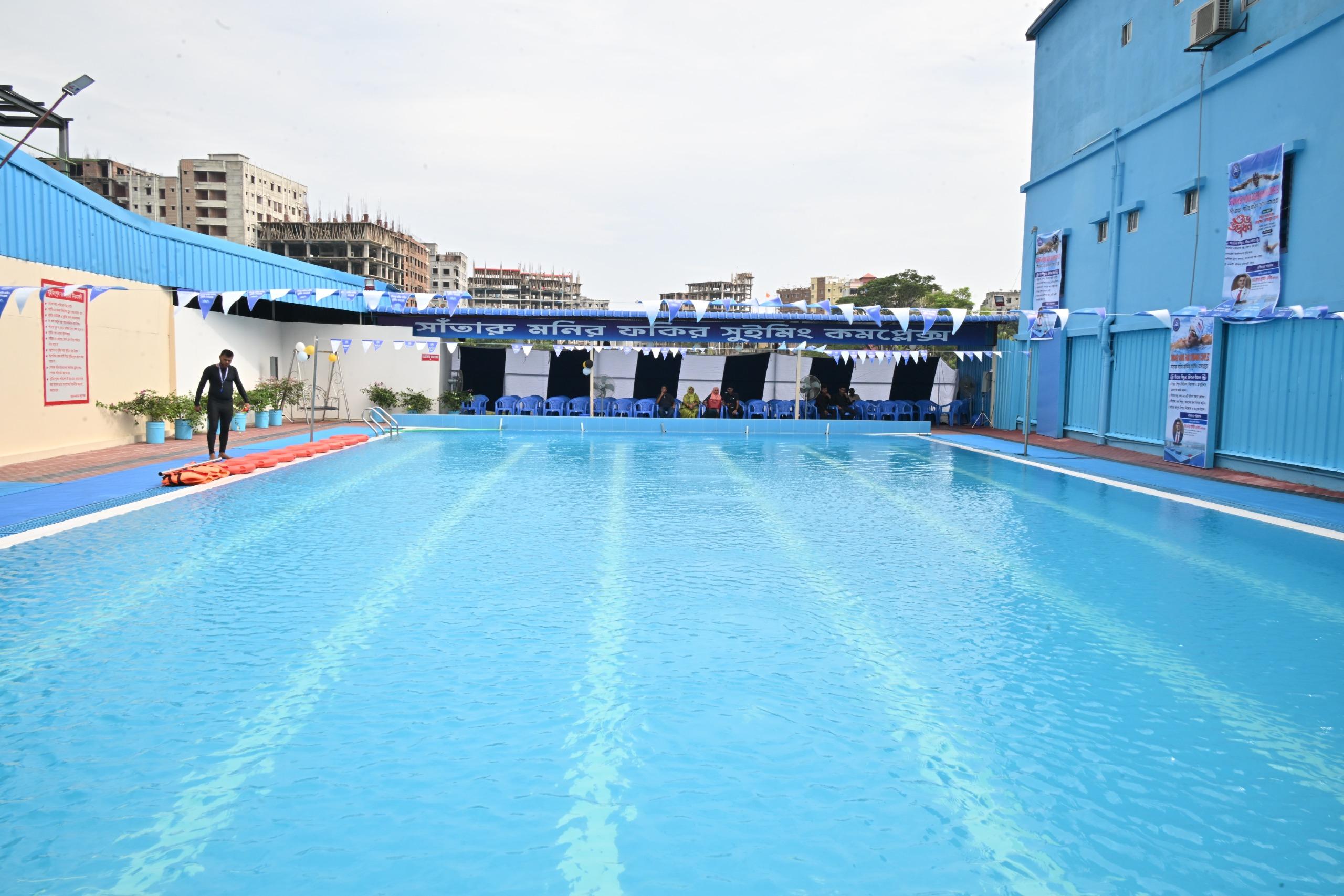 A lifeguard walks the deck of the empty outdoor pool before opening, bunting strung overhead and guests seated along the far wall.