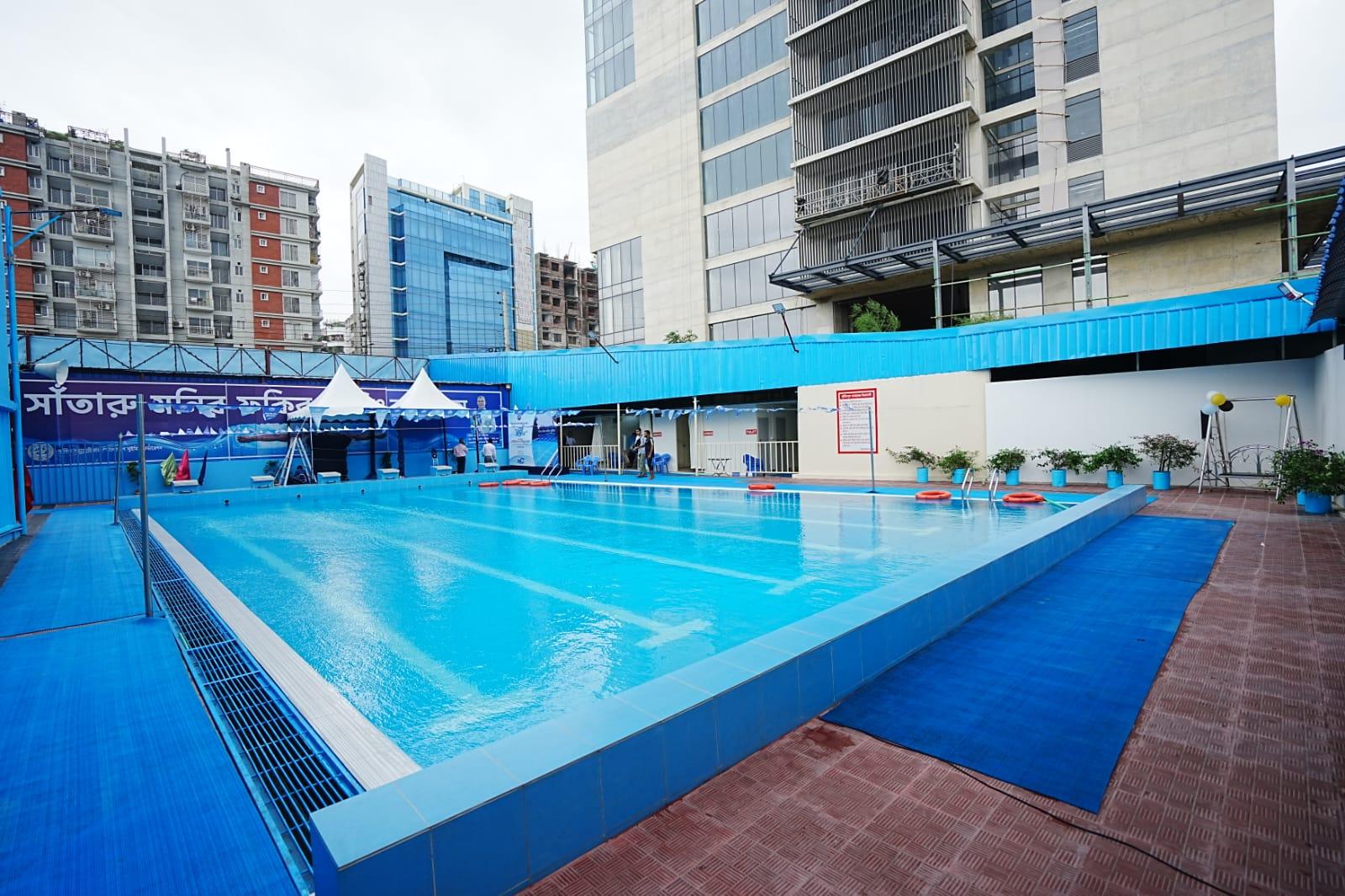 A wide-angle view of the pool deck with rescue rings lined along the lane rope, blue-tiled surrounds, and the city skyline behind.