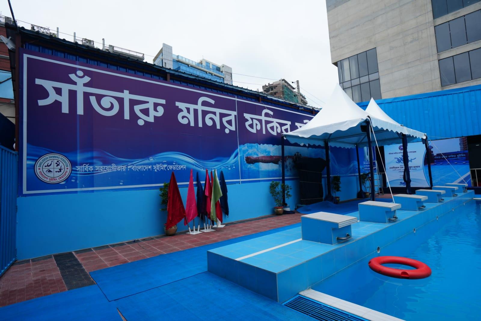 Six starting blocks lined along the pool edge beside red event flags and a Bangladesh Swimming Federation banner, ahead of the ribbon-cutting.