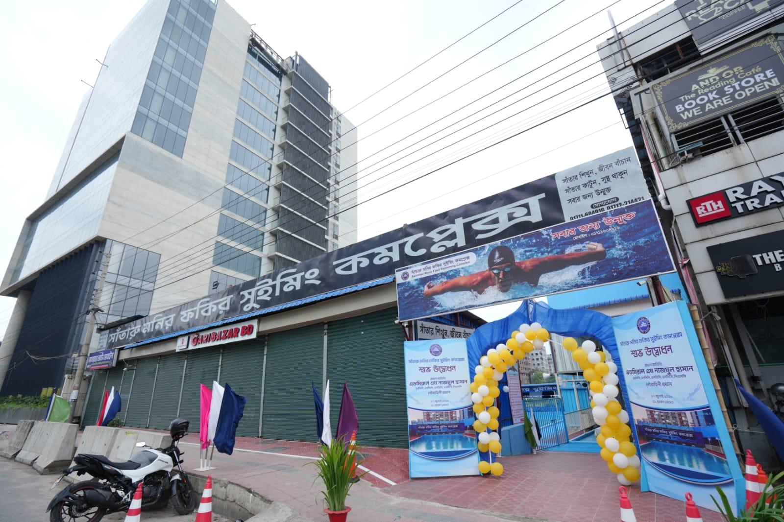 A wide street view of the swimming complex's building facade and rooftop entrance, framed by Dhaka's high-rises under construction.